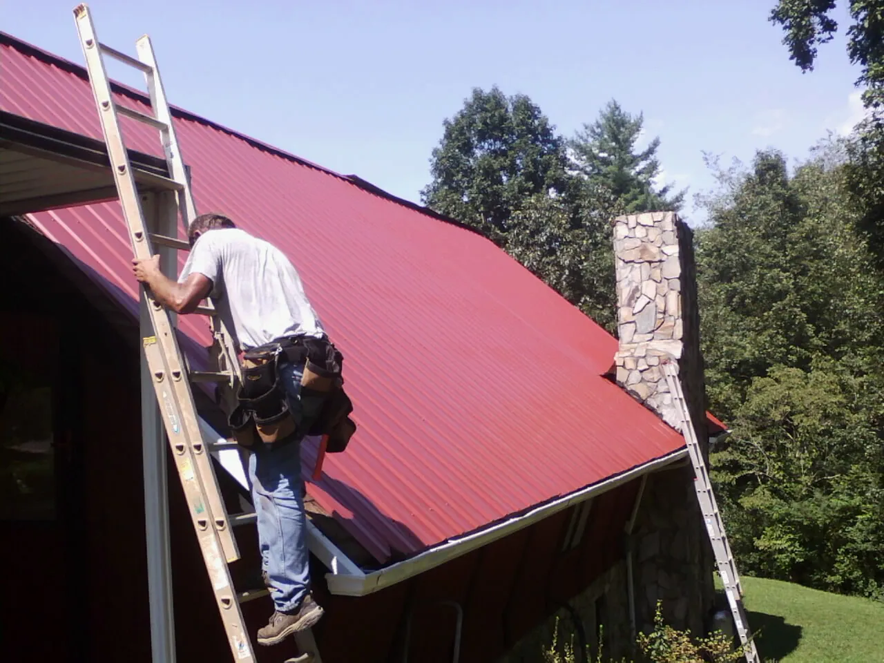 Roofer on a ladder beside a red metal roof and stone chimney — Soffit & Fascia Repair by Roofing Brothers in Saint Leo